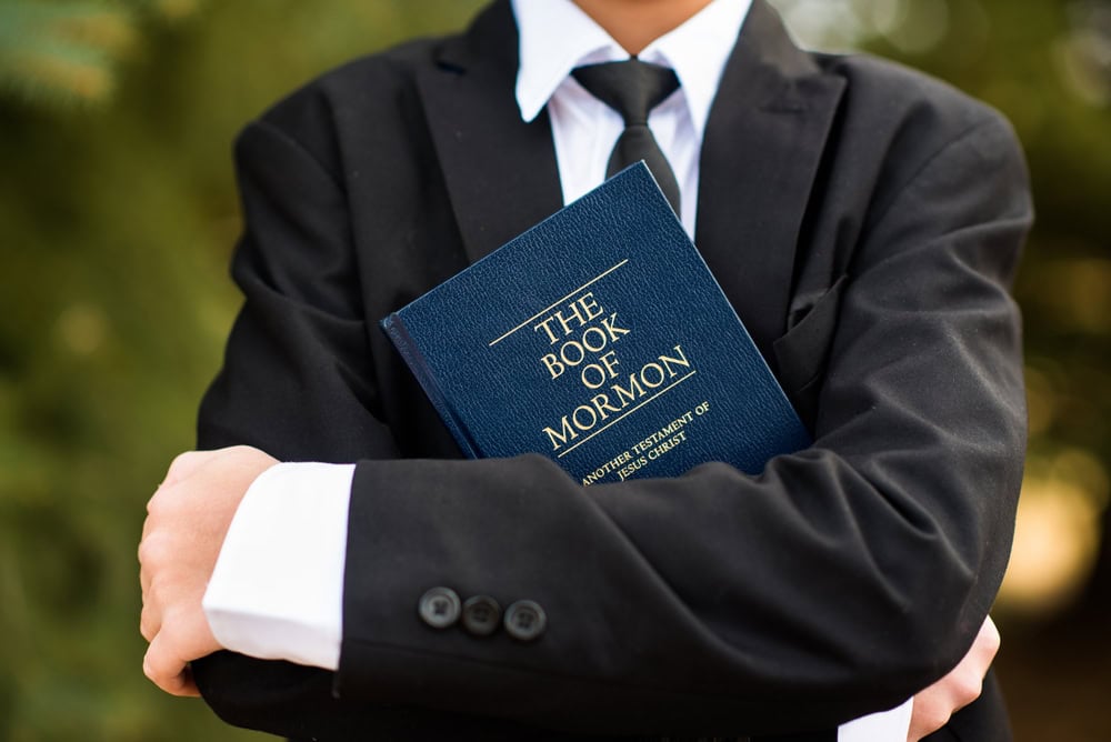 Person in a black suit holding a copy of the Book of Mormon against their chest.