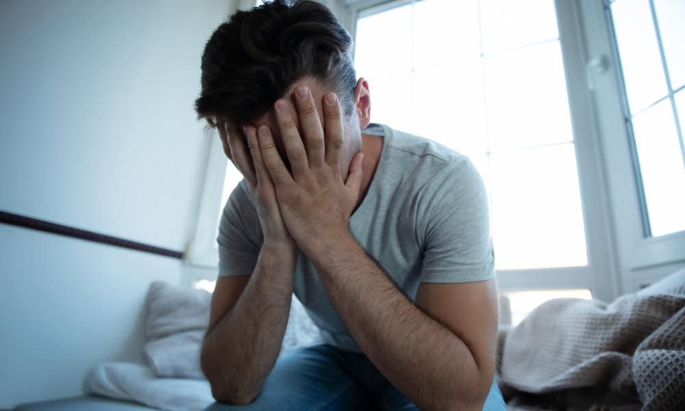 Distressed man sitting on a bed with his face buried in his hands, in a sunlit room, indicative of an LDS sexual abuse survivor.