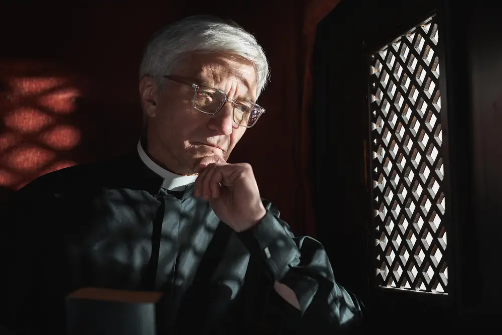 An anonymous priest sits behind the grid of a confessional window looking pensive.