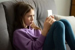 Young girl sitting on a couch wearing a purple hoodie, focused on a smartphone in her hands, representing concerns about online safety for children