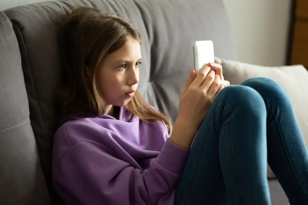 Young girl sitting on a couch wearing a purple hoodie, focused on a smartphone in her hands, representing concerns about online safety for children