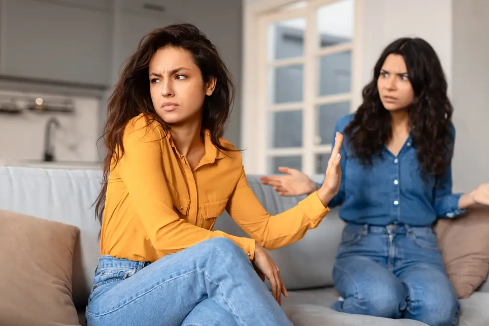 Two women sitting on a couch, one turning away with her hand raised while the other looks upset, illustrating mistakes to avoid when supporting a survivor.