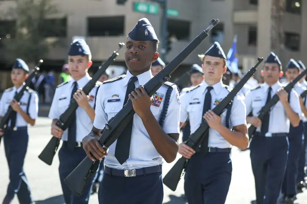 High school JROTC cadets in light blue uniforms march in formation during a parade, each carrying a drill rifle