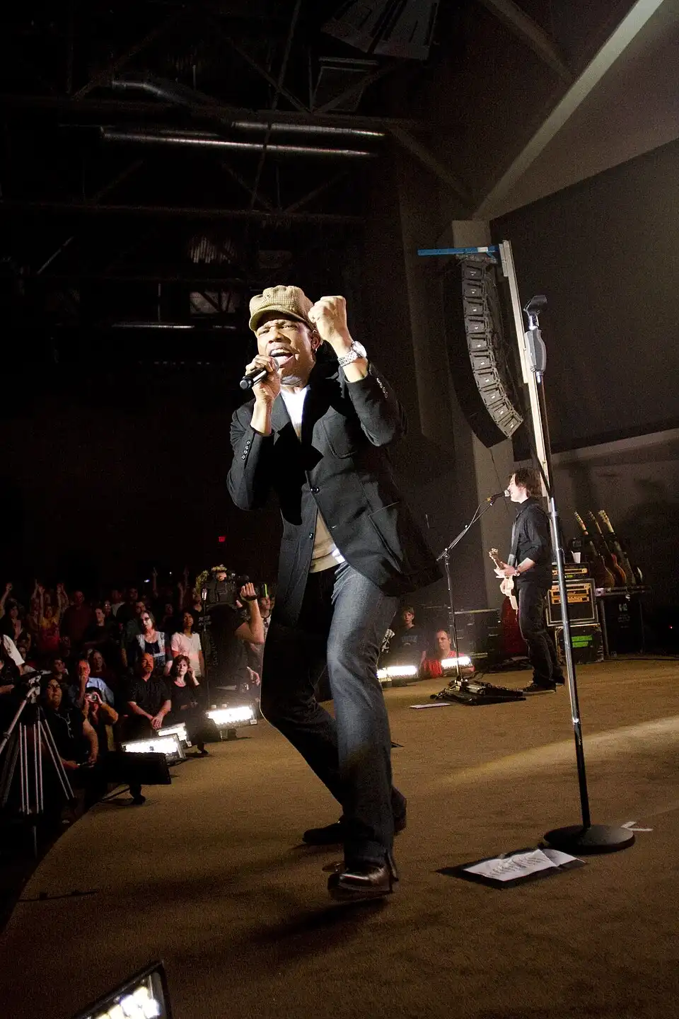 Michael Tait performing on stage with a microphone, wearing a blazer and hat, with a band and cheering audience in the background.