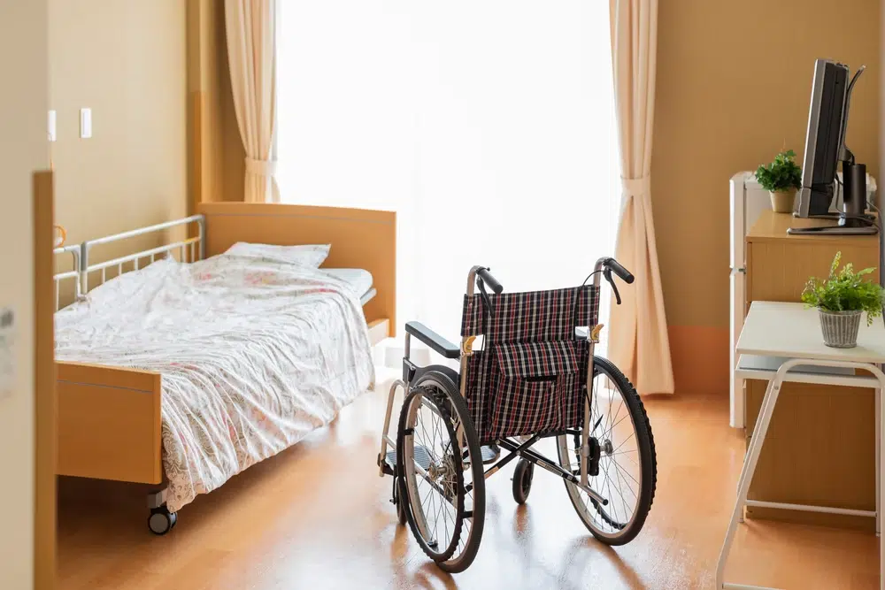 Empty wheelchair in a nursing home bedroom with natural light and medical furniture.