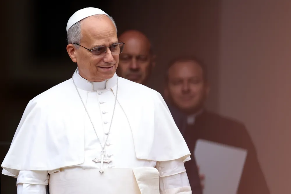 Pope Leo smiles in his white papal robes during his first public appearance, with clergy members standing in the background