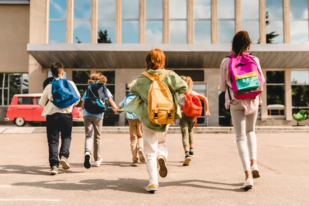 Group of school-aged children wearing backpacks walking toward the entrance of a school building on a sunny day, viewed from behind.