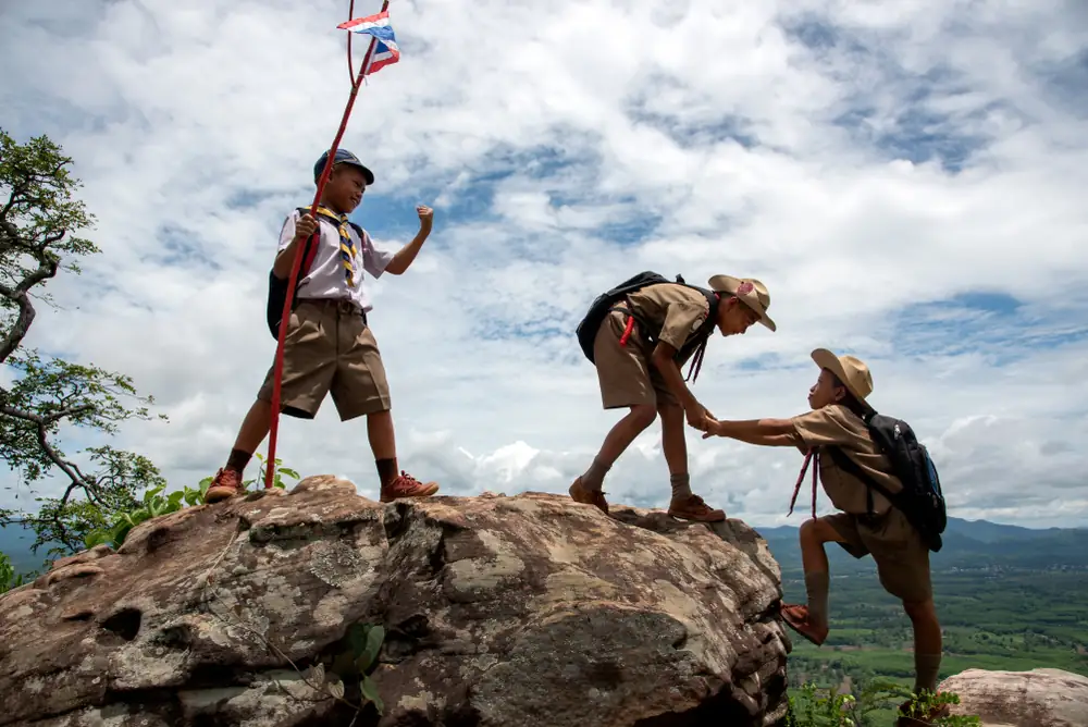 Three scouts climb a mountain, one raises a flag while another helps a friend up the rocky peak.