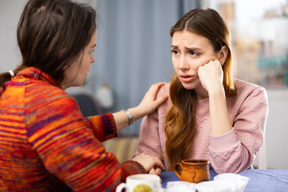 Two women sit at a table, one comforting the other who appears distressed and emotional.