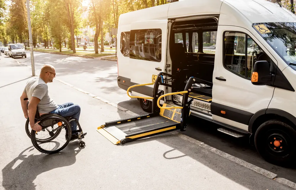 Man in wheelchair preparing to use a ramp lift on an accessible van parked on a sunny street.