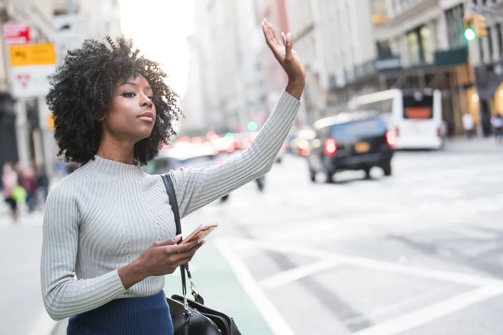Young woman standing on a busy city street, holding her phone and raising her hand to hail a rideshare.