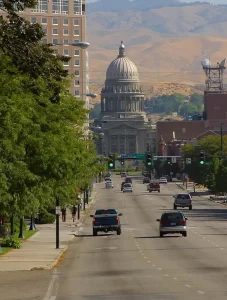 The Idaho State Capitol building in Boise, framed by downtown streets, trees, and cars with rolling hills visible in the background.