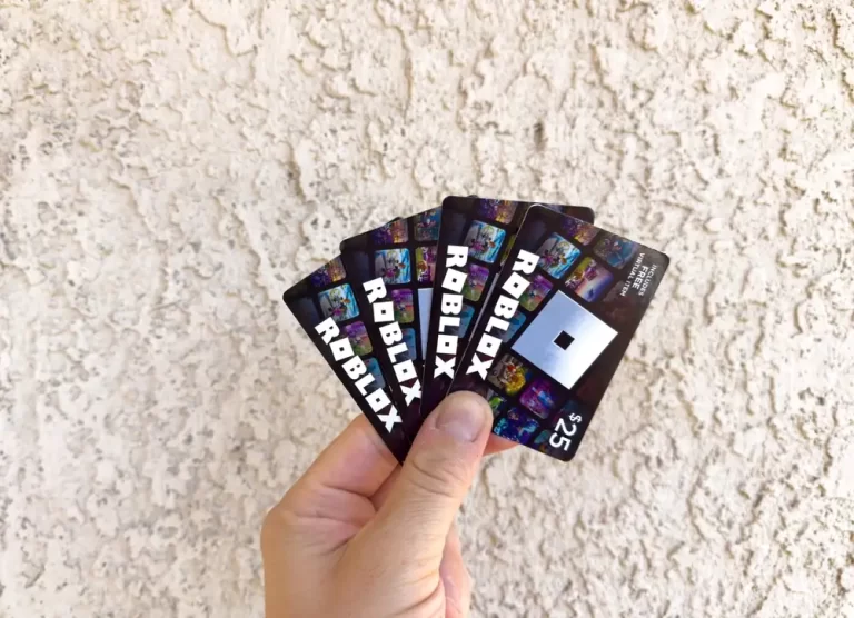 A person holds several $25 Roblox gift cards against a beige textured wall.