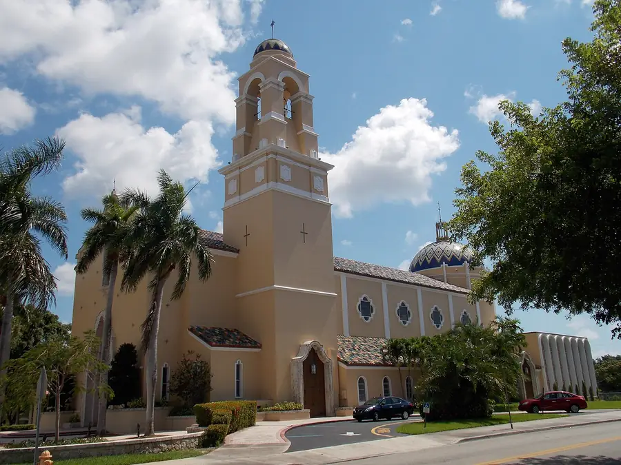 The Cathedral of Saint Mary in Miami, Florida, featuring its yellow exterior, bell tower, palm trees, and a dome against a bright blue sky with clouds.