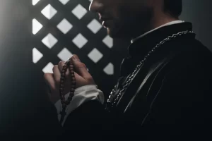 A Catholic priest holding rosary beads while hearing confession inside a dimly lit confessional