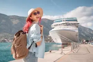 Survivors Rights | Cruise Ship Sexual Assault Lawsuits: The Hidden Contract Clause That Denies Justice A smiling woman with pink hair, sunglasses, and a backpack stands on a pier beside a large cruise ship under a sunny sky with mountains in the background