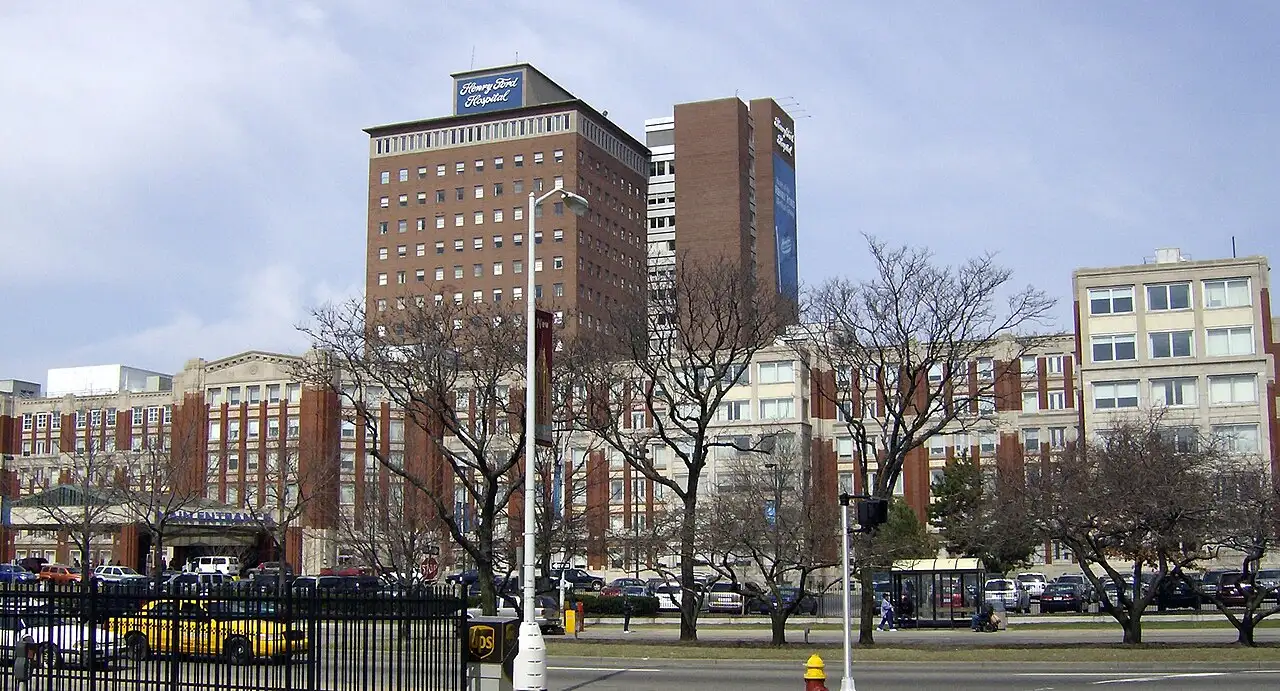 Henry Ford Hospital in Detroit, Michigan, a major medical center with multiple large brick and concrete buildings shown from the street view with cars and trees in front.