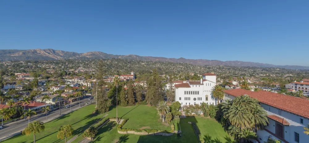 Scenic aerial view of Montecito and Santa Barbara, California, showing Spanish-style buildings, palm trees, and the Santa Ynez Mountains in the background.