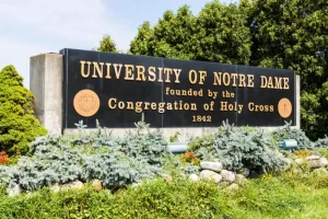Entrance sign of the University of Notre Dame, founded by the Congregation of Holy Cross in 1842, surrounded by greenery and landscaping.