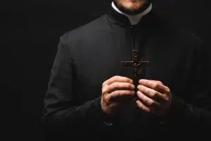 A priest wearing a black clerical robe holds a crucifix with both hands against a dark background.