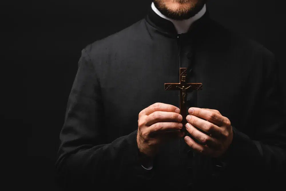 A priest wearing a black clerical robe holds a crucifix with both hands against a dark background.