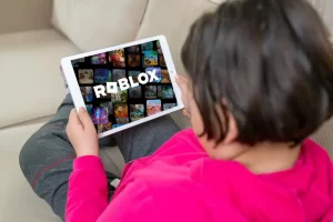 A child sitting on a couch holds a tablet displaying the Roblox game interface, highlighting growing concerns over online child safety on gaming platforms.