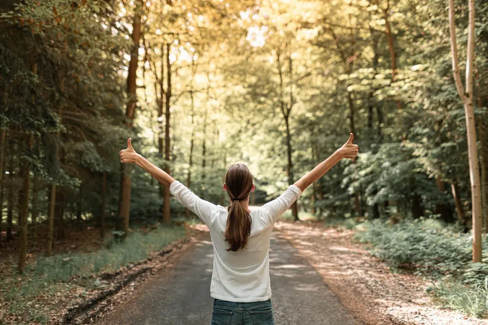 Woman standing on a sunlit forest path with arms raised and thumbs up, symbolizing healing, empowerment, and emotional renewal.
