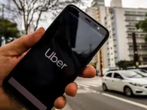 A person holds a smartphone displaying the Uber logo on-screen while standing on a city street with cars and tall buildings in the background