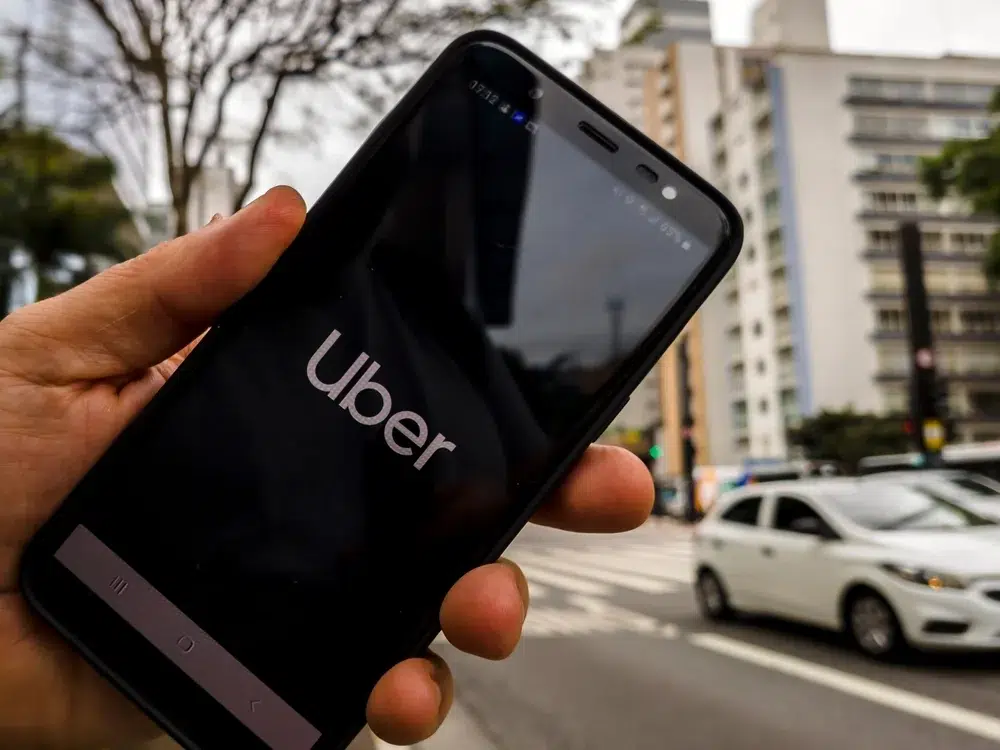 uber-app-on-phone A person holds a smartphone displaying the Uber logo on-screen while standing on a city street with cars and tall buildings in the background