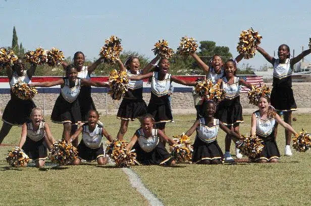 Youth cheerleading team performing a routine outdoors with gold and black pom poms, smiling and posing in coordinated uniforms on a grassy field.