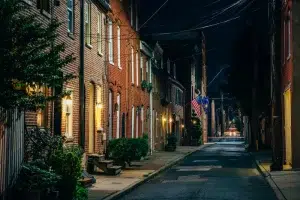 Nighttime view of a narrow residential street lined with historic brick rowhomes, warm porch lights, potted plants, American flags, and overhead utility lines.