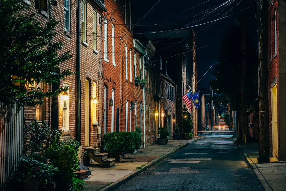 Nighttime view of a narrow residential street lined with historic brick rowhomes, warm porch lights, potted plants, American flags, and overhead utility lines.