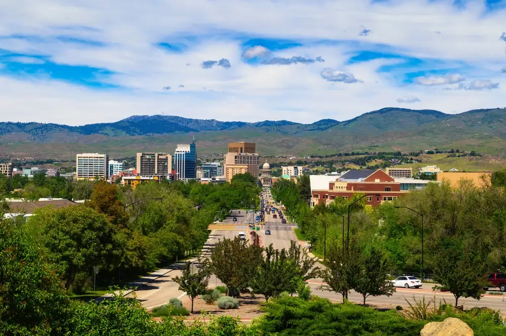 Skyline view of Boise, Idaho with downtown buildings, tree-lined streets, and surrounding foothills under a partly cloudy sky.