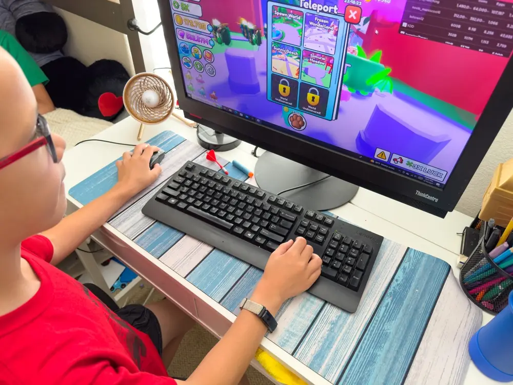 A child wearing a red shirt plays Roblox on a desktop computer at home, with colorful game graphics visible on the screen.