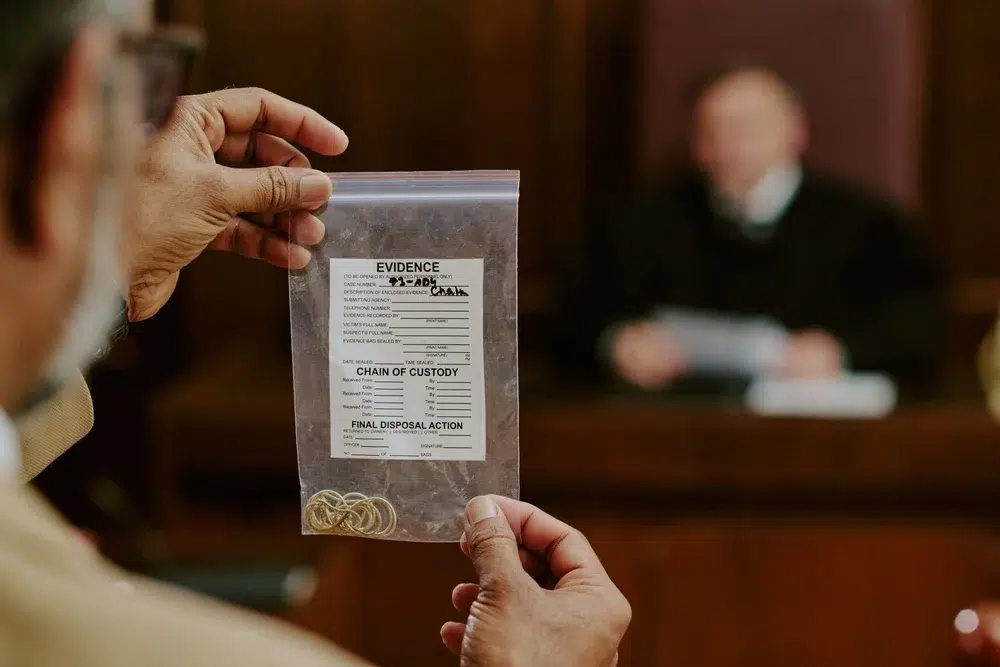 Close-up of a person holding a sealed evidence bag labeled “Chain of Custody” in a courtroom, with a judge blurred in the background.