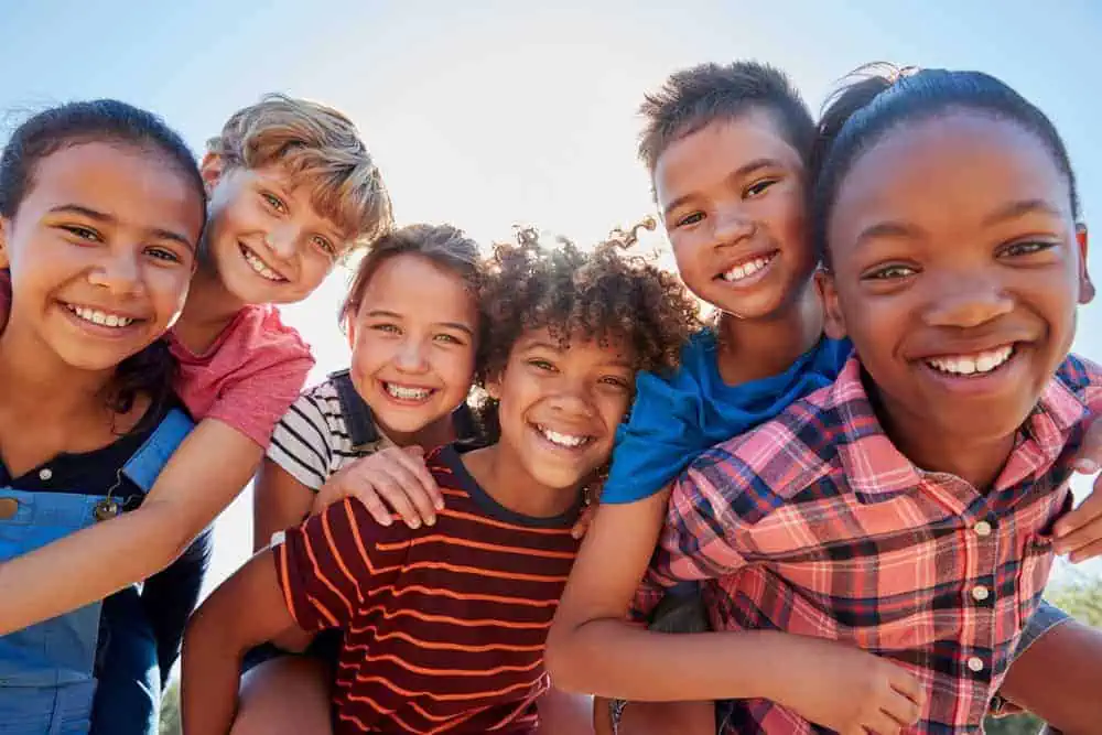 Group of six diverse, smiling children standing outdoors in a close, cheerful pose on a sunny day, symbolizing friendship and childhood joy.