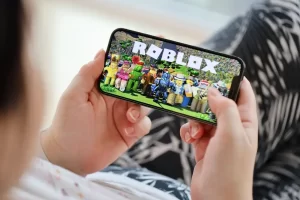 A child holding a smartphone and playing Roblox, displaying colorful block-style avatars on the screen.