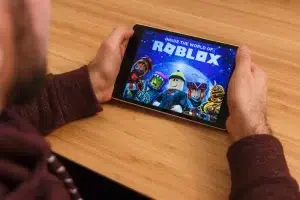 A person holding a tablet and viewing the Roblox game home screen featuring several animated Roblox characters against a space themed background.