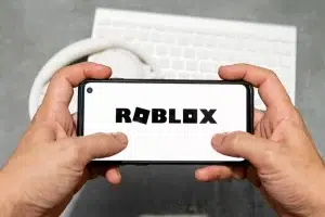 A person holding a smartphone displaying the Roblox logo on the screen, with a white keyboard and headphones in the background.