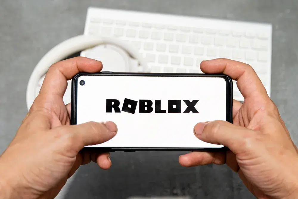A person holding a smartphone displaying the Roblox logo on the screen, with a white keyboard and headphones in the background.