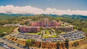 Aerial view of Tripler Army Medical Center in Oahu, Hawaii, showing the large pink hospital complex surrounded by parking areas and lush green mountains.