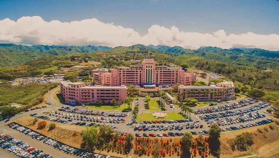 Aerial view of Tripler Army Medical Center in Oahu, Hawaii, showing the large pink hospital complex surrounded by parking areas and lush green mountains.