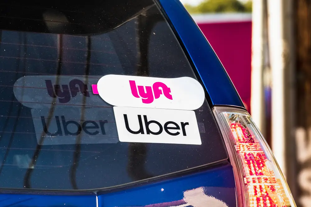 Rear window of a blue car displaying both Uber and Lyft decals, representing a vehicle used for rideshare driving.