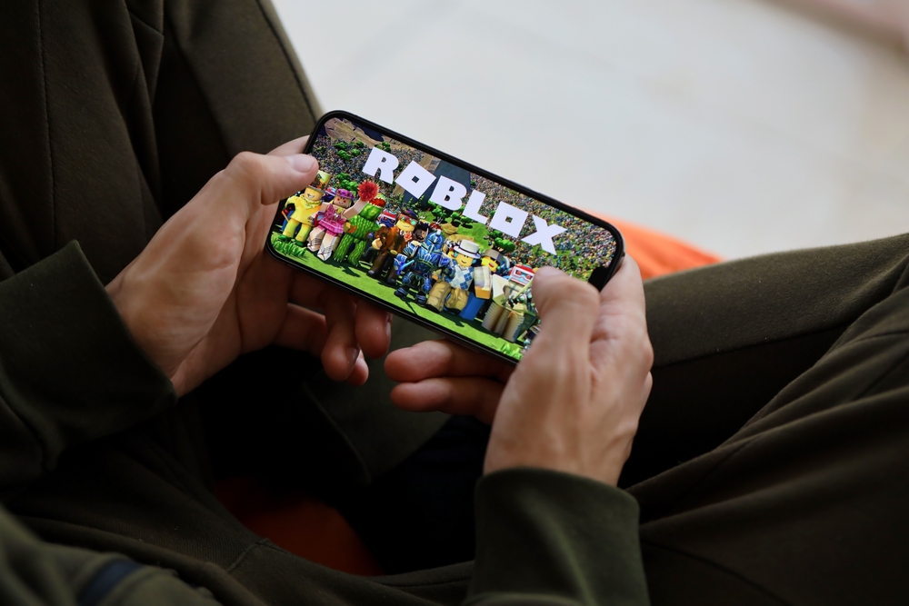 A person holding a smartphone displaying the Roblox game screen with animated characters visible on the device.