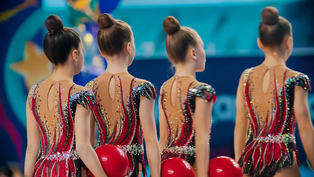 Young rhythmic gymnasts standing in a row during competition, holding red balls and wearing matching sequined leotards.