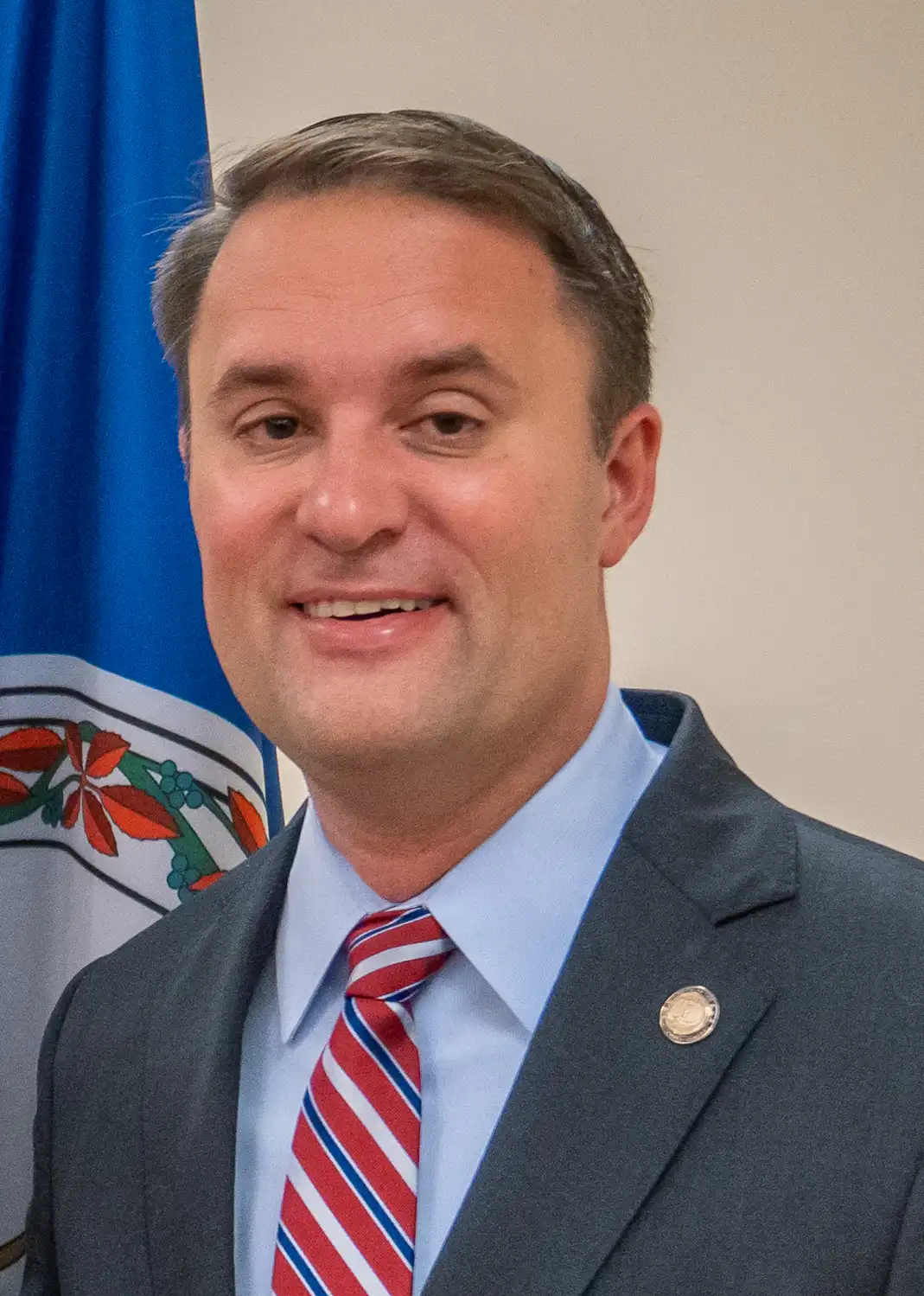 Virginia Attorney General Jason Miyares standing in front of a Virginia state flag, wearing a dark suit and red striped tie.