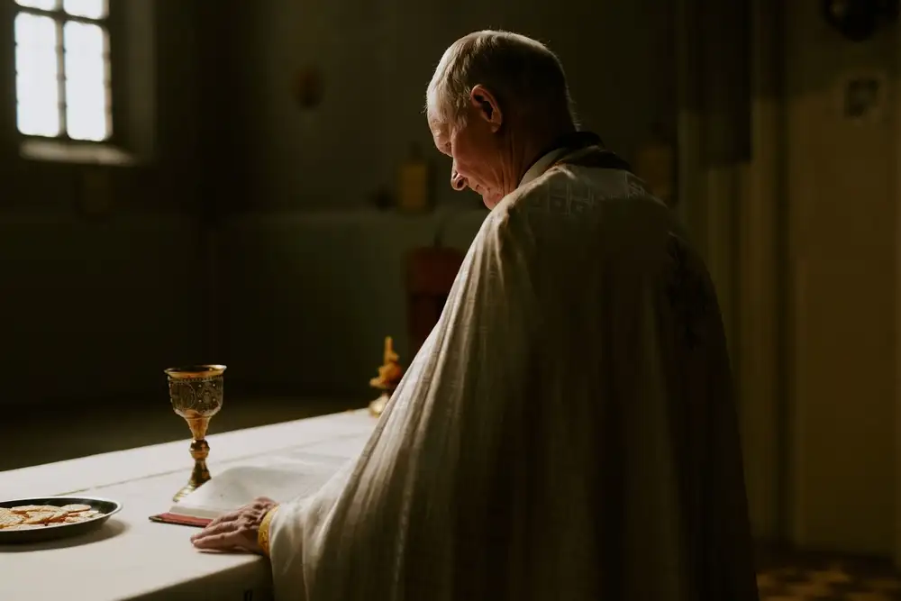 A priest standing at an altar with his head bowed, illuminated by soft light during a religious service.