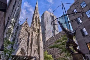 St. Patrick’s Cathedral in New York City with the Atlas statue in the foreground and Midtown Manhattan buildings in the background.