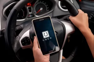 Person holding a smartphone displaying the Uber app logo while sitting in the driver’s seat of a car with hands on the steering wheel.
