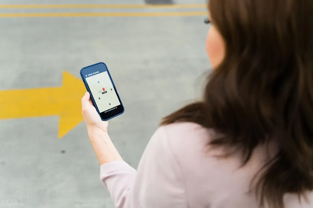 Woman holding a smartphone displaying a rideshare app while standing in a parking area, viewed from behind, illustrating rideshare use and passenger safety concerns.